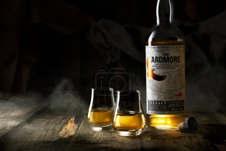 Photo for The Ardmore Whiskey in a bottle with glasses in which alcohol is poured standing on a wooden table surrounded by smoke and fabric background - Royalty Free Image