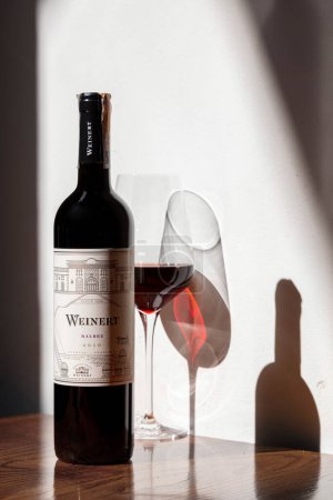 Foto de Una botella de vino tinto weinert malbec 2010 se encuentra sobre una mesa marrón oscuro. Muy cerca se encuentra un frasco de vidrio transparente con vino fresco. A la izquierda, la luz del sol brilla y una sombra cae a la derecha sobre una pared de piedra blanca. - Imagen libre de derechos