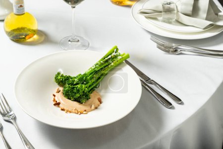 Téléchargez les photos : Choux de brocoli grillés avec sauce aux noix et cacahuètes dans une assiette en céramique légère. Le plat se tient sur une nappe légère, à côté d'un verre de vin blanc et de couverts. - en image libre de droit