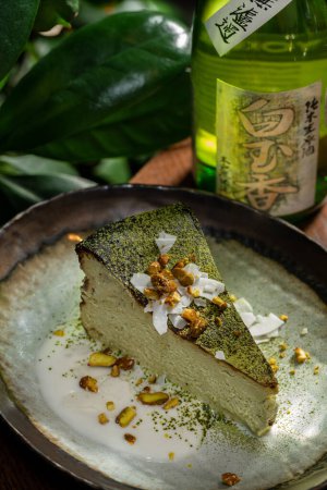 Téléchargez les photos : Gâteau au fromage Matcha aux pistaches et copeaux de noix de coco garni de poudre de thé vert sur une assiette en céramique. A proximité se trouve un verre et une bouteille de vin japonais, et des plantes. - en image libre de droit