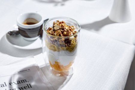 Photo for Ice cream with pineapple, pistachios, muesli, passionfruit, seeds and cheese in a glass on a white table with a cup of coffee - Royalty Free Image