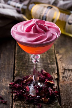 Foto de Un cóctel con helado rosa en un vaso de pie sobre una mesa de madera con hibisco seco, una botella de vino y un paño en el fondo - Imagen libre de derechos