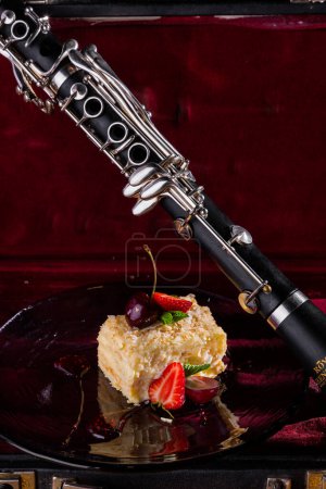 Foto de Un trozo de pastel con crema, migas, mermelada, uvas, fresas, cerezas y menta en un plato que se encuentra sobre la mesa con un oboe en el fondo - Imagen libre de derechos