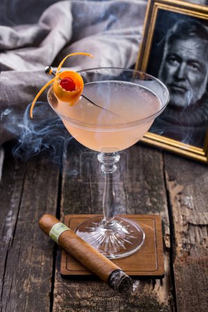 Photo for A cocktail in a glass with orange peel standing on a wooden stand with a photograph book, a smoking cigar and a cloth in the background - Royalty Free Image