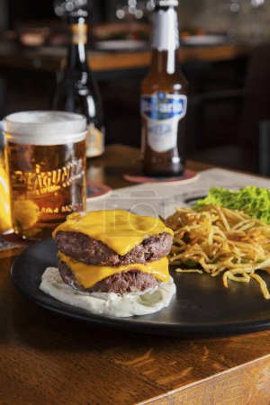 Foto de Dos chuletas de cerdo frito con queso, salsa y palitos de batata en un plato que se encuentra sobre la mesa con un vaso de cerveza y botellas en el fondo - Imagen libre de derechos
