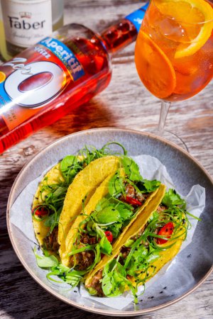 Foto de Tacos con carne, albahaca, perejil, semillas de sésamo, chuka, ensalada en un plato sobre una mesa de madera con un vaso y una botella de aperol y una rebanada de naranja - Imagen libre de derechos