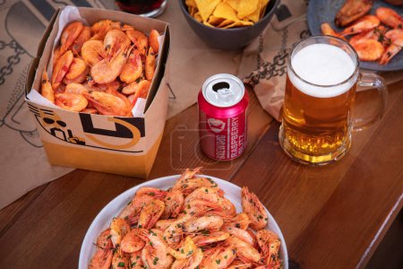 Téléchargez les photos : Crevettes bouillies dans une boîte en carton se tient sur une table avec des crevettes dans un bol, un verre de bière, des copeaux de maïs dans une assiette et une boîte de bière en métal ouvert - en image libre de droit