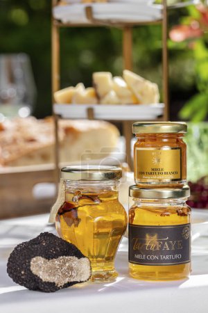 Photo for Honey in jars with truffle mushroom inside. Jars of honey are on the table on a white tablecloth. Behind you can see the cheese platter, different breads, a plate of berries and green plants. - Royalty Free Image