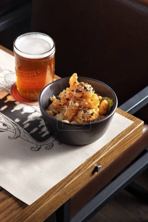 Foto de Nuggets de pollo con semillas de sésamo y salsa en un plato que se encuentra en el borde de la mesa con un vaso de cerveza en el fondo de una silla - Imagen libre de derechos