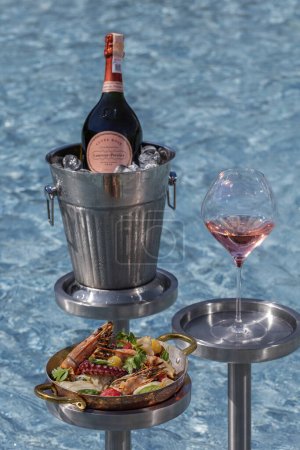 Photo for Assorted grilled seafood lies in a bronze bowl with handles. The plate stands on a metal stand, next to it stands a stand with a glass of wine and a bucket of sparkling wine. coasters are in the pool. - Royalty Free Image