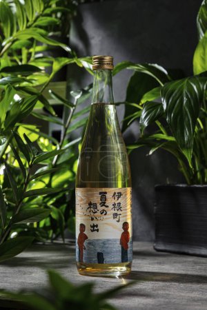 Photo for A bottle of Japanese rice vodka - sake. Sake stands on a gray stone background, tropical plants and a backdrop of gray stone background are arranged around. Nearby stands a wine, transparent glass on a high leg with white wine. - Royalty Free Image
