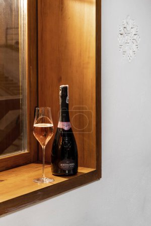 Foto de Botella y una copa de Bortolomiol Filanda Rose Brut ros parada de vino espumoso en una mesa de madera contra una pared de piedra clara con adornos ucranianos y marcos de ventanas de madera. - Imagen libre de derechos