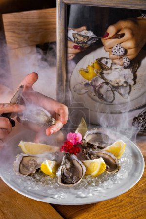 Foto de Las manos humanas pelan una ostra que yace en un plato con hielo, limón y pétalos de flores que está sobre la mesa sobre el fondo de un marco fotográfico - Imagen libre de derechos