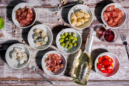 Foto de Snacks for wine, prosciutto, Parmesan cheese, blue cheese, peppers, sausages, olives in plates on the table with a bottle of wine, cutlery and glasses of drinks - Imagen libre de derechos