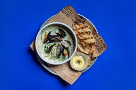 Foto de Creamy soup with seafood, squid, salmon, mussels, potatoes and chuka seaweed. The soup is poured into a round, ceramic bowl and stands on a newspaper sheet. Nearby are two toasted French baguettes and a sauceboat of lemon cream sauce. The food is in - Imagen libre de derechos