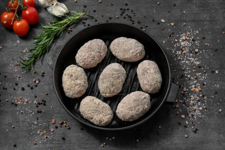 Photo for Turkey cutlets in flour breading. They lie on a black cast iron pan. The dishes stand on a dark stone background, spices and herbs are scattered around. - Royalty Free Image