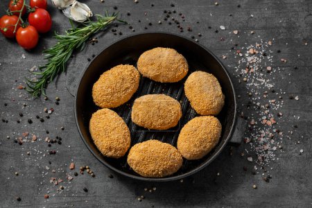 Photo for Turkey cutlets in breadcrumbs. They lie on a black cast iron pan. The dishes stand on a dark stone background, spices and herbs are scattered around. - Royalty Free Image
