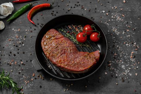 Photo for Veal tenderloin in marinade. It lies on a black cast iron pan. The dishes stand on a dark stone background, spices and herbs are scattered around. - Royalty Free Image