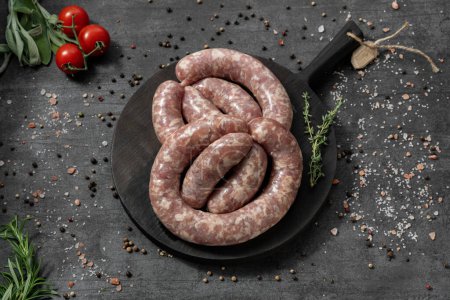 Photo for Pork sausages with spicy herbs. They lie on a black, wooden, round board. The dishes stand on a dark stone background, spices and herbs are scattered around. - Royalty Free Image