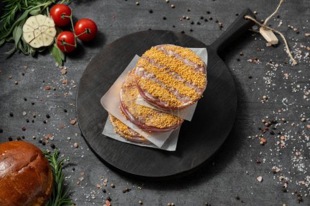 Photo for Beef cutlets for burgers with breading in bacon. They lie on a black, wooden, round board. The dishes stand on a dark stone background, spices and herbs are scattered around. - Royalty Free Image