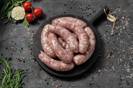 Photo for Pork sausages with pieces of paprika and spicy herbs. They lie on a black, wooden, round board. The dishes stand on a dark stone background, spices and herbs are scattered around. - Royalty Free Image