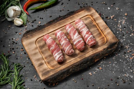 Foto de Kebab de carne envuelto en tocino. Se encuentra en una tabla de madera ligera. Los platos están sobre un fondo de piedra oscura, especias y hierbas se dispersan alrededor. - Imagen libre de derechos