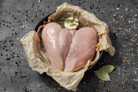 Photo for Chicken fillet on parchment in a cast iron pan. The dishes stand on a dark stone background, spices and herbs are scattered around. - Royalty Free Image