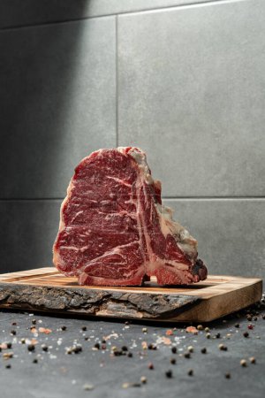 Photo for T-bone steak on a light wooden board, standing on a dark stone background, with salt and pepper mixed with peas scattered around. - Royalty Free Image