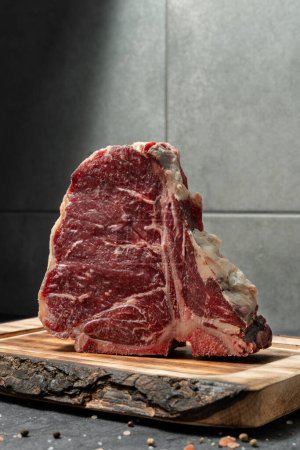 Téléchargez les photos : T-bone steak sur une planche de bois clair, debout sur un fond de pierre sombre, avec du sel et du poivre mélangés avec des pois dispersés autour. - en image libre de droit