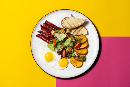 Foto de Desayuno inglés de dos huevos fritos, salchichas, papas fritas, tostadas y ensalada. La comida se encuentra en un plato de cerámica blanca sobre un fondo de color papel. - Imagen libre de derechos