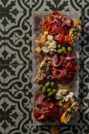 Photo for On a wooden board are sliced cheese and meat snacks for wine. Brie, Camembert, Parmesan and salami cheeses, prosciutto and cured meats. Next to the olives and bruschetta. The board rests on a tile with a pattern. - Royalty Free Image
