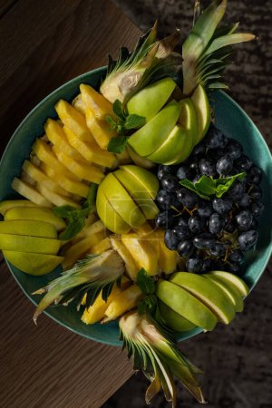 Téléchargez les photos : L'ananas, les pommes et les raisins rouges coupés en morceaux se trouvent dans un bol en céramique profonde. Placez le piala sur une table en bois. - en image libre de droit