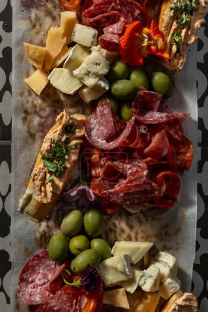 Photo for On a wooden board are sliced cheese and meat snacks for wine. Brie, Camembert, Parmesan and salami cheeses, prosciutto and cured meats. Next to the olives and bruschetta. The board rests on a tile with a pattern. - Royalty Free Image