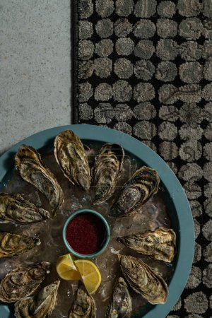 Téléchargez les photos : Douze huîtres sur un oreiller de glace dans une assiette ronde bleue. Au centre, placer un bol avec sauce framboise et quartiers de citron. - en image libre de droit