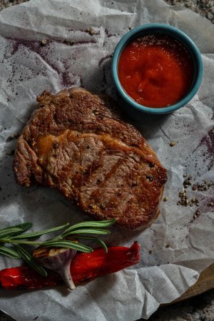 Téléchargez les photos : Le steak de porc repose sur du parchemin blanc sur une planche de bois. Un bol de sauce tomate et un brin de romarin sont à proximité.. - en image libre de droit