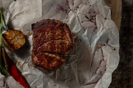 Foto de Filet mignon steak se encuentra en el pergamino blanco sobre una tabla de madera. Cerca están los chiles rojos, el ajo asado y una ramita de romero. - Imagen libre de derechos