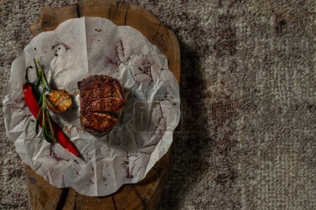 Photo for Filet mignon steak lies on white parchment on a wooden board. Nearby are red chili peppers, roasted garlic and a sprig of rosemary. - Royalty Free Image