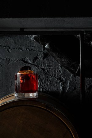 Téléchargez les photos : Cocktail avec mezcal et vermouth, liqueur et poire fumée, garni de frites d'orange dans un verre à l'ancienne. Tenez un verre avec un cocktail sur un tonneau en bois. - en image libre de droit