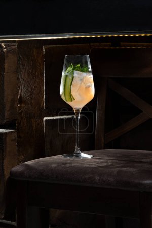 Téléchargez les photos : Cocktail avec gin blanc et rose, jus de citron, sirop de sureau et une tranche de concombre et une feuille de menthe. Boire dans un verre sur une tige haute sur une surface en cuir. - en image libre de droit