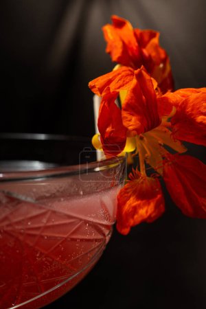 Téléchargez les photos : Cocktail avec vermouth et gin, purée de fraises, amers à la rhubarbe et liqueur de sureau. Décoré avec une fleur dans un verre transparent à tige. Prenez un cocktail sur une chaise brune. - en image libre de droit