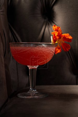 Téléchargez les photos : Cocktail avec vermouth et gin, purée de fraises, amers à la rhubarbe et liqueur de sureau. Décoré avec une fleur dans un verre transparent à tige. Prenez un cocktail sur une chaise brune. - en image libre de droit