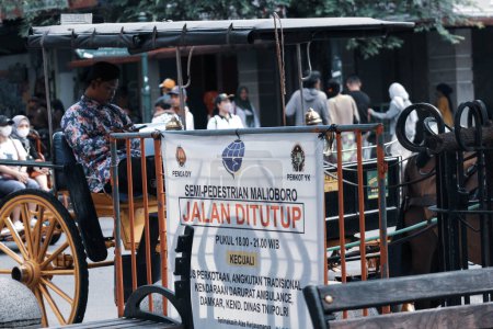 Téléchargez les photos : Yogyakarta, Indonésie en octobre 2022. Un conducteur de chariot et son chariot s'arrêtent devant une bannière qui lit Street Closed. La rue semi-piétonne Malioboro est fermée de 18.00 à 20.00 WIB. - en image libre de droit