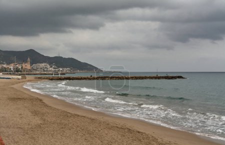 Photo for Empty beach Sitges, Catalunya ,Spain in winter with the overcast sky and a leaden day with a gentle swell and its typical church in the background with empty space for text. - Royalty Free Image