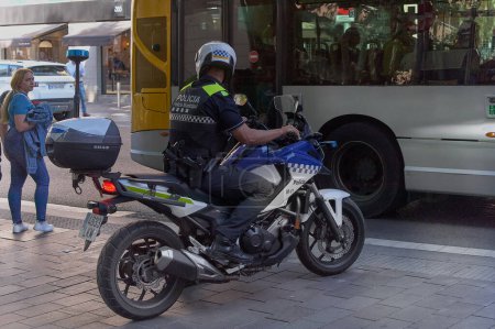 Téléchargez les photos : Gava - 30 mai 2023 : Un motard de la police municipale de Gava conduit sa moto, surveillé par un citoyen alors qu'un bus de passagers passe. - en image libre de droit