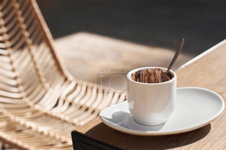 White cup with traces of chocolate, illuminated by sunlight, on a wooden table next to a woven chair.