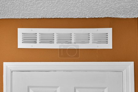 Image of a white ventilation grill embedded in a white wall, with a light brown border and a textured white ceiling. Perfect for projects related to interior architecture or climate control.