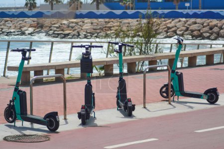 Three electric scooters parked along the railing of a boardwalk, with the ocean and rocks in the background, ready to be rented and explore the coast