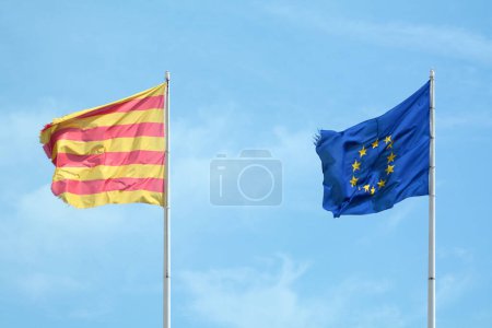 L'image capture un moment symbolique où le drapeau de l'Union européenne et le drapeau de Catalogne flottent ensemble contre un ciel bleu clair, représentant l'unité dans la diversité.