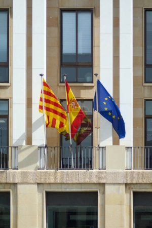 The Spanish national flag, the Catalan Senyera, and the European Union flag wave side by side, symbolizing unity amidst diversity against a modern architectural backdrop.
