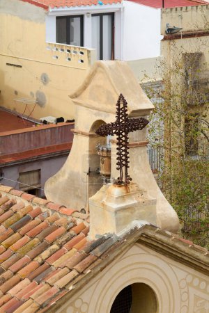 Foto de Una cruz de metal ornamentado se destaca sobre un telón de fondo de edificios urbanos que se alza sobre un antiguo techo con azulejos rojos y una estructura arquitectónica detallada.. - Imagen libre de derechos